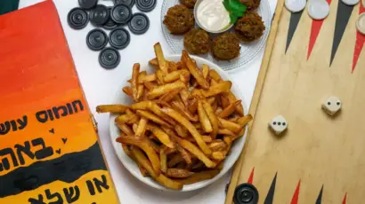 צלחת מלאה בצ'יפס פריך לצד מסבחה וחומוס, עם משחק דמקה ברקע. חומוס הבוטקה בגליל.